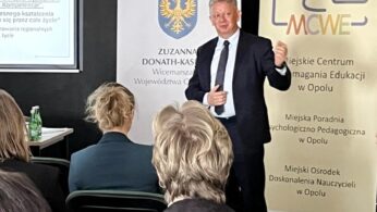 Mężczyzna w garniturze prowadzi prezentację dla publiczności w sali konferencyjnej. Za nim znajdują się dwa banery z logo i polskim tekstem oraz ekran projektora wyświetlający częściowo widoczny slajd. Trzech słuchaczy siedzi naprzeciwko niego.