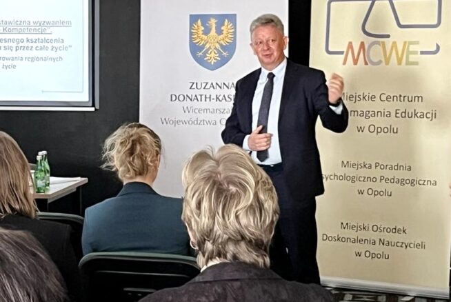 Mężczyzna w garniturze prowadzi prezentację dla publiczności w sali konferencyjnej. Za nim znajdują się dwa banery z logo i polskim tekstem oraz ekran projektora wyświetlający częściowo widoczny slajd. Trzech słuchaczy siedzi naprzeciwko niego.