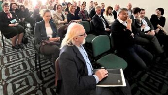 Grupa ludzi siedzi w rzędach krzeseł w sali konferencyjnej, uważnie patrząc przed siebie. Większość z nich ubrana jest w stroje biznesowe. Pokój ma wzorzystą wykładzinę, kremowe ściany i duże okna z tyłu wpuszczające jasne światło słoneczne.