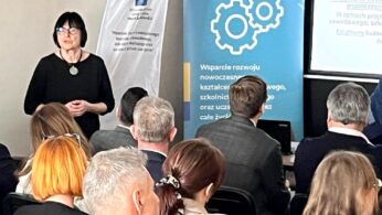 Kobieta z krótkimi ciemnymi włosami i w okularach prowadzi prezentację dla siedzącej publiczności w sali konferencyjnej. Banery z logo i tekstem w języku polskim są wyświetlane za nią obok ekranu z częściowo widocznym wyświetlanym slajdem.