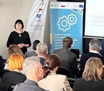Kobieta z krótkimi ciemnymi włosami i w okularach prowadzi prezentację dla siedzącej publiczności w sali konferencyjnej. Banery z logo i tekstem w języku polskim są wyświetlane za nią obok ekranu z częściowo widocznym wyświetlanym slajdem.