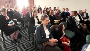 Grupa dorosłych, zarówno mężczyzn, jak i kobiet, siedzi uważnie w kilku rzędach krzeseł w dobrze oświetlonej sali konferencyjnej. Większość z nich ubrana jest w stroje biznesowe lub formalne. Światło słoneczne wpada przez okno z tyłu, tworząc jasną, przyjazną atmosferę.