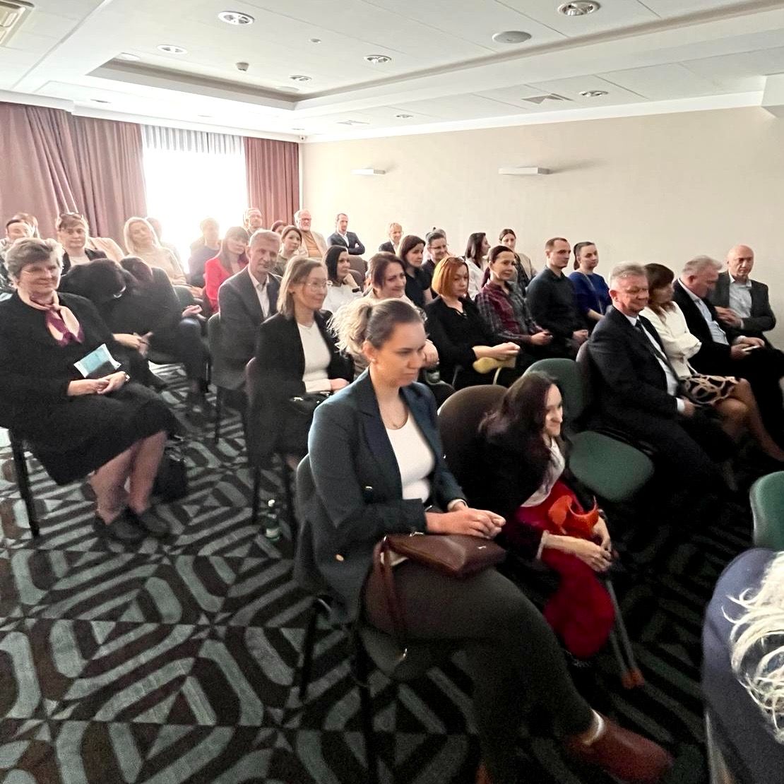 Grupa dorosłych, zarówno mężczyzn, jak i kobiet, siedzi uważnie w kilku rzędach krzeseł w dobrze oświetlonej sali konferencyjnej. Większość z nich ubrana jest w stroje biznesowe lub formalne. Światło słoneczne wpada przez okno z tyłu, tworząc jasną, przyjazną atmosferę.
