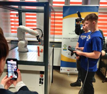 Dwóch nastolatków w niebieskich koszulkach obsługuje ramię robota wewnątrz szklanej obudowy za pomocą kontrolera. Jedna osoba filmuje kamerą, a druga robi zdjęcie smartfonem. Żółto-biały baner stoi w tle przy oknach z czerwonymi roletami.