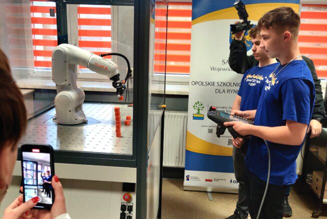 Dwóch nastolatków w niebieskich koszulkach obsługuje ramię robota wewnątrz szklanej obudowy za pomocą kontrolera. Jedna osoba filmuje kamerą, a druga robi zdjęcie smartfonem. Żółto-biały baner stoi w tle przy oknach z czerwonymi roletami.