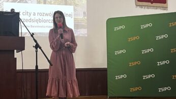 Kobieta w różowej wzorzystej sukience i czarnych butach stoi na drewnianej podłodze, trzymając mikrofon. Za nią znajduje się ekran projekcyjny z prezentacją, zielony baner ZSIPO i polskie godło na ścianie.