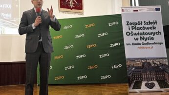 Mężczyzna w garniturze przemawia do mikrofonu na scenie. Za nim znajduje się zielone tło z logo ZSIPO, polski orzeł na ścianie i pionowy baner Zespołu Szkół i Placówek Oświatowych w Nysie. Sceneria wydaje się być formalnym wydarzeniem wewnątrz budynku.