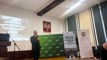 Mężczyzna w garniturze przemawia do mikrofonu na scenie w pokoju wyłożonym drewnianymi panelami. Za nim znajdują się banery, zielone tło z logo ZSPO, ekran projekcyjny z polskim tekstem i godło Polski na ścianie. Członkowie publiczności są widoczni na pierwszym planie.