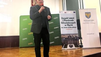 Starszy mężczyzna w czarnym garniturze stoi na drewnianej scenie i mówi do mikrofonu. Za nim znajdują się zielone, białe i niebieskie banery z polskim tekstem oraz godło polskiej flagi na ścianie. Wydaje się, że jest to impreza szkolna lub środowiskowa.