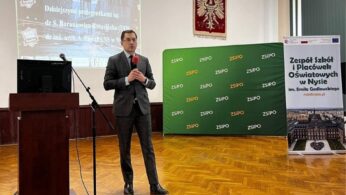 Mężczyzna w garniturze trzyma mikrofon i przemawia na scenie w klasie z projektorem wyświetlającym tekst, zielony baner i stojący plakat o Zespole Szkół Oświatowych w Nysie. Logotypy i informacje o finansowaniu są wyświetlane w prawym dolnym rogu.
