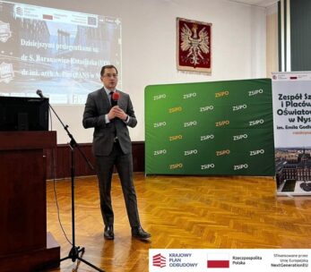 Mężczyzna w garniturze trzyma mikrofon i przemawia na scenie w klasie z projektorem wyświetlającym tekst, zielony baner i stojący plakat o Zespole Szkół Oświatowych w Nysie. Logotypy i informacje o finansowaniu są wyświetlane w prawym dolnym rogu.