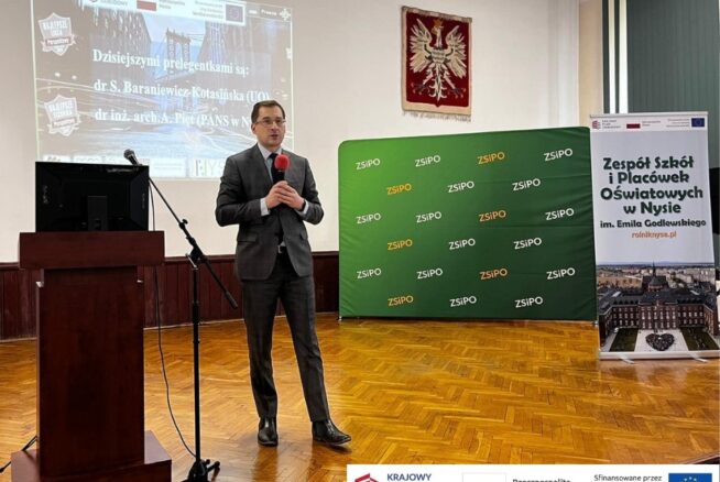 Mężczyzna w garniturze stoi na drewnianej scenie trzymając mikrofon, prezentując w sali z zielonym tłem ZSIPO i sztandarem szkoły. W dolnej części obrazu widoczne są logotypy Krajowego Planu Odbudowy, Rzeczpospolitej Polskiej i Unii Europejskiej.
