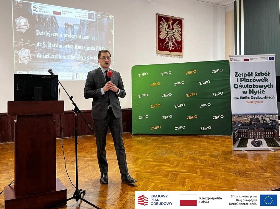Mężczyzna w garniturze trzyma mikrofon i przemawia na scenie w klasie z projektorem wyświetlającym tekst, zielony baner i stojący plakat o Zespole Szkół Oświatowych w Nysie. Logotypy i informacje o finansowaniu są wyświetlane w prawym dolnym rogu.