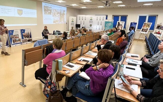 Kobieta stoi i prezentuje slajdy klasie uważnych dorosłych siedzących przy biurkach i robiących notatki. W pokoju znajdują się plakaty, broszury i niebieskie krzesła. Dwie projekcje pokazują logo i tekst na przedniej ścianie. Wygląda to na profesjonalne seminarium.