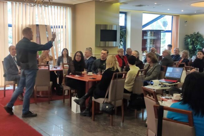 Mężczyzna stoi i przemawia do grupy dorosłych siedzących przy stołach w dobrze oświetlonej sali konferencyjnej. Uczestnicy słuchają uważnie. Na stołach znajduje się projektor, kubki i papiery. Duże okna, żyrandole i rośliny zdobią nowoczesną przestrzeń.