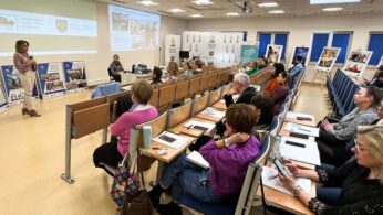 Kobieta stoi z przodu klasy, prowadząc prezentację dla siedzącej publiczności. Uczestnicy robią notatki i patrzą na ekran wyświetlający slajdy. Na ścianach wiszą tablice z plakatami, a pomieszczenie jest jasne dzięki dużym oknom i górnym światłom.