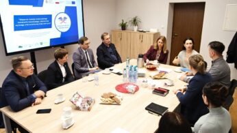 Dziewięć dorosłych osób, pięciu mężczyzn i cztery kobiety, siedzi przy prostokątnym stole w biurowej sali konferencyjnej. Są zaangażowani w dyskusję. Na stole znajdują się napoje, dokumenty i przekąski. Na dużym ekranie wyświetlana jest prezentacja z tekstem i logo w języku polskim.
