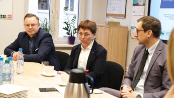 Trzy osoby w strojach biznesowych siedzą przy stole konferencyjnym z butelkami wody, kubkami i termosami. Za nimi znajduje się okno, kalendarz, pliki i ekran prezentacji. Wyglądają na zaangażowanych w profesjonalne spotkanie lub dyskusję.