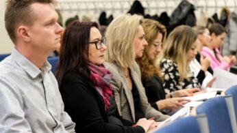 Rząd dorosłych siedzi w klasie lub sali konferencyjnej, uważnie słuchając i robiąc notatki. Niektórzy noszą okulary, a w tle wiszą płaszcze. Otoczenie wydaje się formalne, z naciskiem na naukę lub dyskusję.