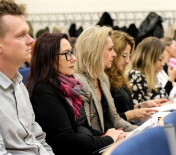 Rząd dorosłych siedzi w klasie lub sali konferencyjnej, uważnie słuchając i robiąc notatki. Niektórzy noszą okulary, a w tle wiszą płaszcze. Otoczenie wydaje się formalne, z naciskiem na naukę lub dyskusję.