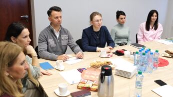 Sześć osób siedzi przy stole z butelkowaną wodą, ciastkami, przekąskami i papierami. Wyglądają na zaangażowanych w spotkanie lub dyskusję. Widoczne są notatniki i długopisy, a atmosfera wygląda na profesjonalną i skupioną.