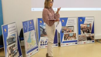 Kobieta we wzorzystej bluzce, beżowych spodniach i brązowych butach stoi i gestykuluje, trzymając tablet przed pięcioma dużymi plakatami na sztalugach, które przedstawiają mapy, zdjęcia i polskie teksty związane z wydarzeniami biznesowymi i edukacyjnymi.