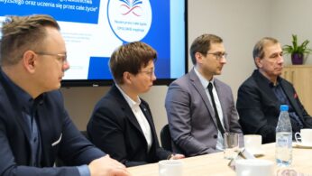 Cztery osoby w formalnych strojach siedzą przy stole konferencyjnym z kubkami, butelką wody i papierami. Za nimi niebieski ekran wyświetla polski tekst i logo z otwartą książką, sugerując dyskusję lub spotkanie na temat uczenia się przez całe życie na Opolszczyźnie.