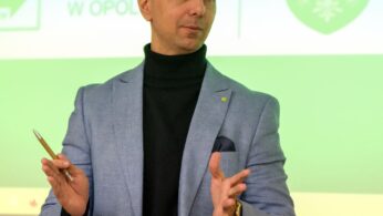 Mężczyzna z krótkimi ciemnymi włosami, ubrany w jasnoniebieską marynarkę, czarny golf i trzymający długopis, gestykuluje rękami podczas mówienia. Za nim znajduje się zielono-biały slajd prezentacji z herbem i częściowym polskim tekstem.
