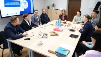 Dziesięć osób ubranych w stroje biznesowe siedzi przy prostokątnym stole konferencyjnym w sali konferencyjnej. Na stole leżą papiery, butelki z wodą, kubki, przekąski i notatniki. Na dużym ekranie wyświetlana jest prezentacja na temat szkoleń i wsparcia zatrudnienia.