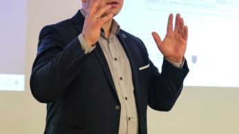 Mężczyzna w ciemnej marynarce i jasnej koszuli przemawia, gestykulując uniesionymi rękami. Nosi okulary i pierścionek, pojawiając się w połowie prezentacji przed wyświetlanym slajdem z tekstem, w dobrze oświetlonym wnętrzu.