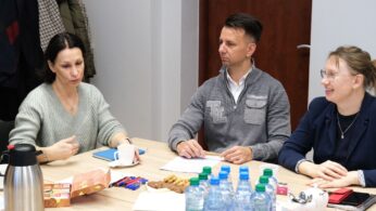 Trzy osoby siedzą przy stole i dyskutują. Kobieta po lewej gestykuluje podczas mówienia, mężczyzna w środku słucha z długopisem i notatnikiem, a kobieta po prawej uśmiecha się. Na stole znajdują się butelki z wodą, ciastka, przekąski, termos i długopisy.