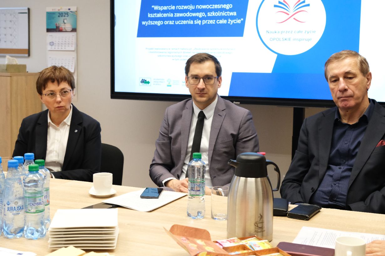 Trzy osoby w strojach biznesowych siedzą przy stole konferencyjnym z butelkami wody, dzbankiem do kawy i papierami. Za nimi znajduje się ekran wyświetlający polski tekst na temat uczenia się przez całe życie i edukacji zawodowej, a na ścianie wisi kalendarz.