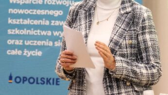 Kobieta z blond włosami spiętymi w wysoki kok, w okularach, białym golfie i kraciastej kurtce, trzyma dokumenty i przemawia. Za nią znajduje się niebieski znak z białymi kołami zębatymi i polskim tekstem, a na pierwszym planie stół z butelkami wody.