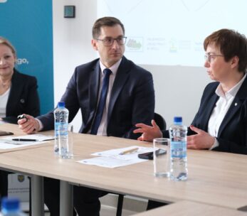 Trzy osoby w strojach biznesowych siedzą przy stole konferencyjnym z dokumentami i butelkami wody. Kobieta po prawej stronie mówi, gestykulując rękami, podczas gdy mężczyzna w środku i kobieta po lewej stronie słuchają. W tle widoczny jest ekran projekcyjny.