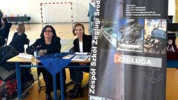 Dwie kobiety siedzą przy stoliku z broszurami w sali gimnastycznej. Obok nich znajduje się pionowy baner 