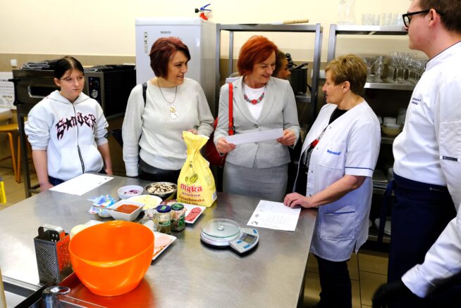 Pięć osób stoi wokół stołu kuchennego ze stali nierdzewnej ze składnikami do pieczenia, wagą i przepisami. Trzy kobiety rozmawiają z kobietą w białym fartuchu; jedna młoda osoba stoi z boku. W tle półki ze szklankami i przyborami kuchennymi.
