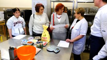 Pięć dorosłych osób stoi wokół stołu kuchennego ze stali nierdzewnej, na którym znajdują się składniki takie jak mąka, grzyby, ser i miska do mieszania. Jedna kobieta w białym fartuchu trzyma dokumenty, sugerując lekcję gotowania lub demonstrację. W tle znajdują się półki ze szklankami i talerzami.