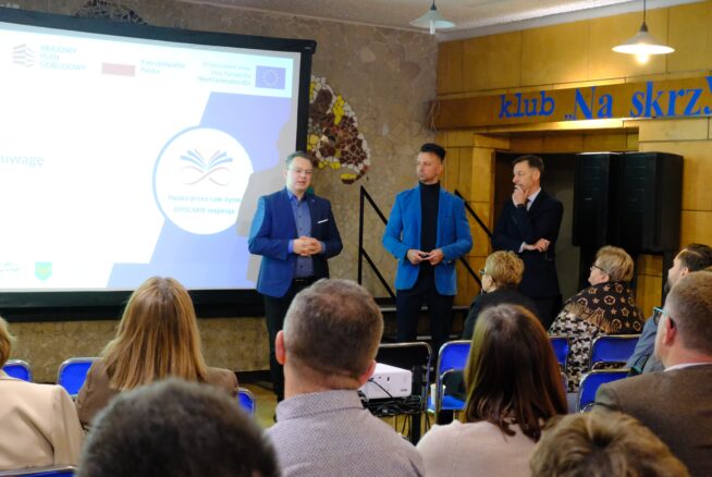 Trzech mężczyzn stoi przed siedzącą publicznością w pokoju o ścianach wyłożonych drewnianymi panelami. Przemawiają w pobliżu dużego ekranu projektora wyświetlającego slajd. Nad i za nimi widoczny jest niebieski znak i logo UE. Publiczność słucha uważnie.