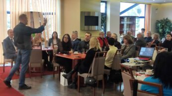 Mężczyzna stoi i gestykuluje, przemawiając do grupy osób siedzących przy stołach w dobrze oświetlonej sali konferencyjnej. Uczestnicy słuchają uważnie, z notatnikami, kubkami i projektorem na pobliskim stole, co sugeruje ustawienie biznesowe lub warsztatowe.