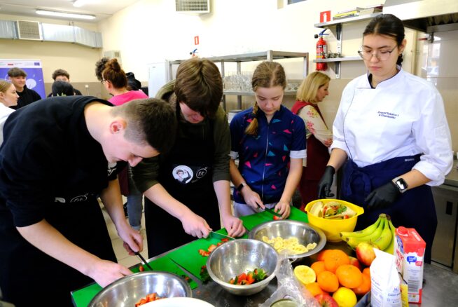 Czworo nastolatków przygotowuje jedzenie przy blacie kuchennym, krojąc warzywa i owoce na zielonych deskach do krojenia. Kobieta w fartuchu szefa kuchni i rękawiczkach nadzoruje pracę. Na blacie stoją miski, banany i pomarańcze. W tle widać inne osoby.