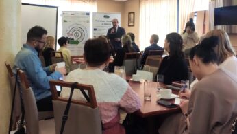 Grupa ludzi siedzi przy stołach w sali konferencyjnej, słuchając mężczyzny przemawiającego z przodu. Widoczny jest ekran projektora i kolorowa tablica prezentacyjna. Większość uczestników jest zwrócona do przodu; niektórzy robią notatki. Fotograf stoi w pobliżu dużych okien w tle.