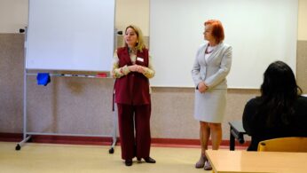Dwie kobiety stoją z przodu klasy, jedna w bordowej kamizelce i spodniach, druga w jasnoszarym garniturze. Stoją twarzą do siedzących uczestników, a za nimi znajduje się tablica i ekran projekcyjny. Otoczenie wydaje się formalne i edukacyjne.