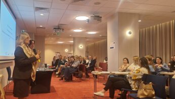 Kobieta w garniturze mówi do mikrofonu z przodu sali konferencyjnej wypełnionej siedzącymi uczestnikami. Obok niej stoi inna kobieta. Na ścianie wyświetlana jest prezentacja. Publiczność słucha uważnie w dobrze oświetlonym, nowoczesnym otoczeniu.
