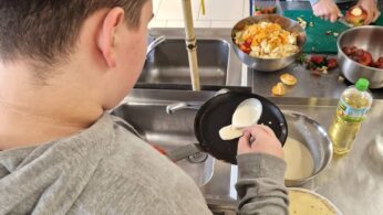 Osoba w szarej bluzie z kapturem wylewa ciasto na małą patelnię nad zlewem kuchennym. Na blacie znajduje się talerz naleśników, miska mieszanych owoców, butelka oleju i inna osoba krojąca owoce na zielonej desce. Kuchnia ma podłogę wyłożoną kafelkami i metalowe urządzenia.