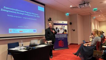 Mężczyzna stoi przed ekranem projekcyjnym wyświetlającym polską prezentację w sali konferencyjnej. Siedzący uczestnicy słuchają. Laptopy i sprzęt leżą na stole. Widoczny jest zwijany baner i nowoczesne lampy sufitowe.