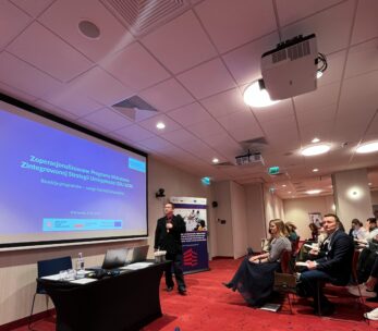 Mężczyzna prowadzi prezentację w nowoczesnej sali konferencyjnej z ekranem wyświetlającym slajd w języku polskim. Siedzący uczestnicy, niektórzy robiący notatki, stoją naprzeciw niego. Pokój ma czerwony dywan, białe ściany i oświetlenie sufitowe. W tle widoczny jest baner i projektor.