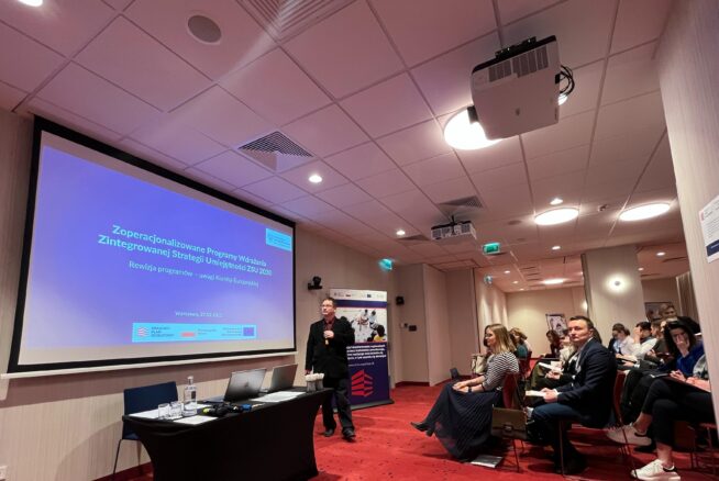 Mężczyzna prowadzi prezentację w nowoczesnej sali konferencyjnej z ekranem wyświetlającym slajd w języku polskim. Siedzący uczestnicy, niektórzy robiący notatki, stoją naprzeciw niego. Pokój ma czerwony dywan, białe ściany i oświetlenie sufitowe. W tle widoczny jest baner i projektor.