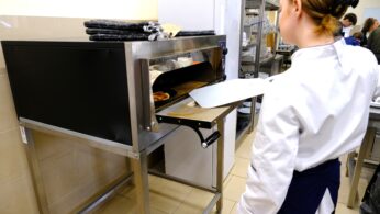 Osoba w białym fartuchu szefa kuchni używa metalowej łopatki do pizzy, aby wyjąć pizzę z pieca ze stali nierdzewnej w komercyjnej kuchni. Rękawice kuchenne są ułożone na wierzchu piekarnika, a w tle widać kilka osób pracujących.