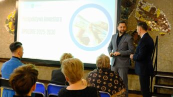 Grupa ludzi obserwuje dwóch mężczyzn w garniturach przemawiających przed ekranem projektora wyświetlającego tytuł 