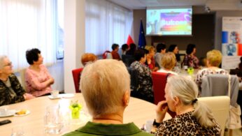 Grupa starszych osób, głównie kobiet, siedzi przy stole konferencyjnym i ogląda prezentację na ekranie, na którym wyświetlane jest słowo 